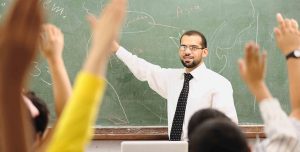teacher at chalkboard; students raising hands to answer a question