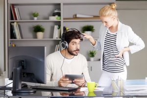 man wearing headphones sits at desk, woman stands by and lifts one ear of the headphones